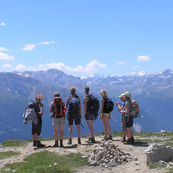 Zes wandelaars met rugzakken staan op een bergpad en kijken uit over een schilderachtig uitzicht op besneeuwde bergtoppen in de verte, onder een strakblauwe hemel.